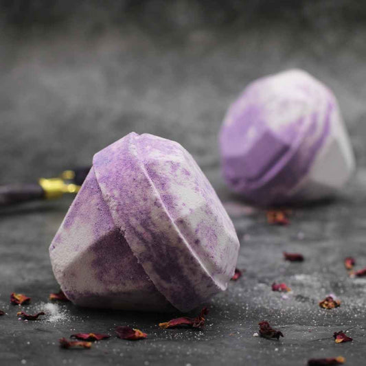 A picture of two luxurious looking purple or lilac diamond shaped bath bombs. The bath bombs are laying on their sides on a dark grey background. Sprinkles of dried red rose petals in the foreground and between the two bath bombs add to the sense of luxury. You can see a blurred partial image of a black crystal facial roller in the background behind the two bath bombs.