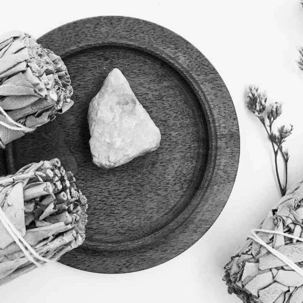 Black and white photo of a plate with the edge of two white sage smudge sticks on the edge of the plate. Next to the smudge sticks on the plate is a crystal. There is another smudge stick on the right of the picture, next to the smudge stick is a sprig of lavender