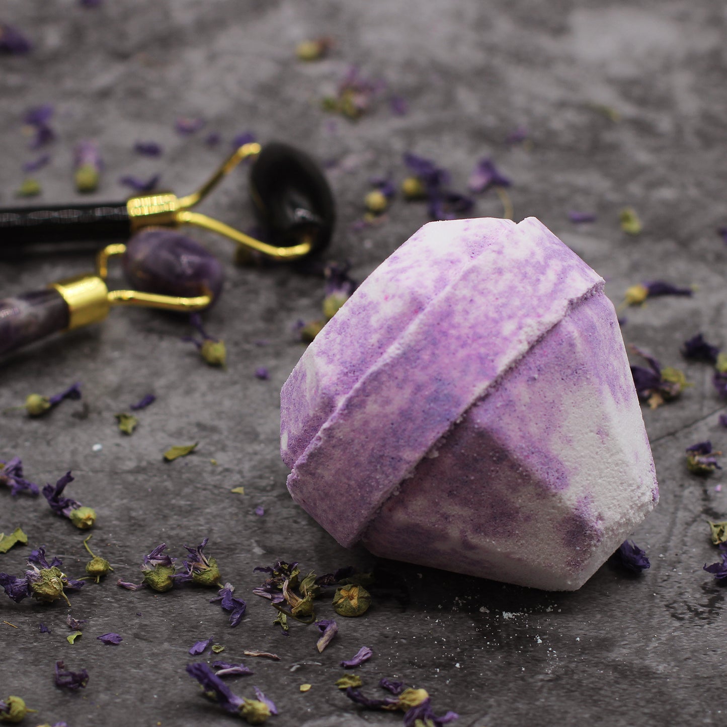 A picture of a luxurious looking purple or lilac diamond shaped bath bomb. The bath bomb is laying on its side on a dark grey background. Sprinkles of dried purple and yellow flower petals in the foreground and between the bath bomb  and two crystal facial rollers, adding to the sense of luxury.