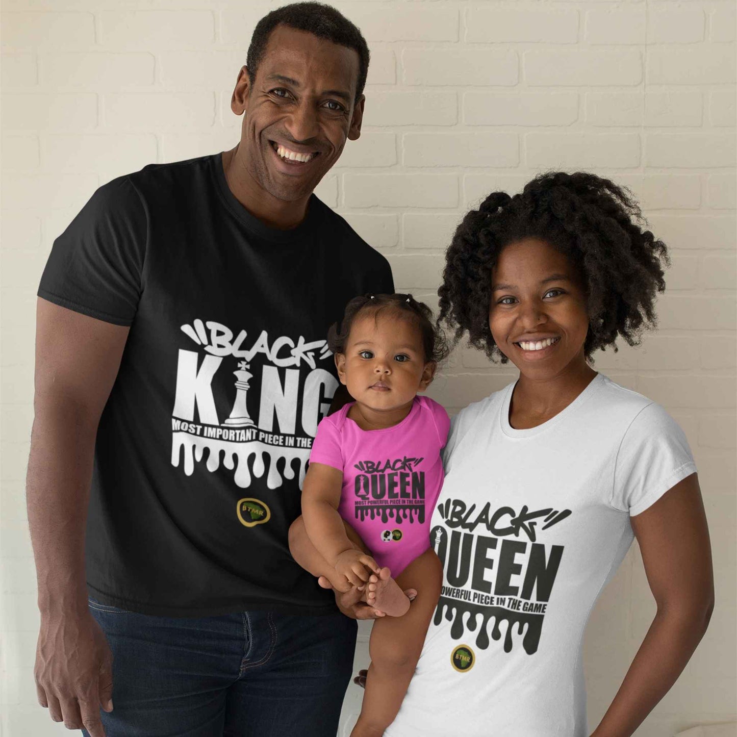 Picture shows a black family, mum, dad and baby wearing their black king and black queen chess statement t shirts. Black woman has on a white fitted t shirt, short sleeves, with Black Queen in black font on the front. She's holding a baby who is wearing a black queen babygrow, they are with the dad who wears a black t shirt, which says Black King the most powerful piece on the board.