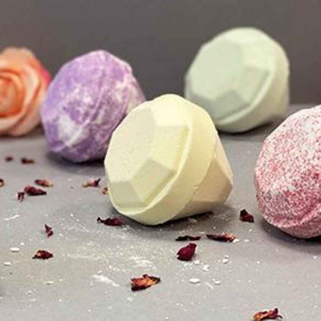 Picture is of a collection of colourful bath bombs against a dark grey background. There are 4 diamond shaped jewel bath bombs laying on their sides. Each bath bomb is a different colour - purple, light yelllow, light green and pink. There is also a peach coloured rose. The picture looks luxurious with sprinkles of dried rose petals in the foreground. The bath bombs look big.
