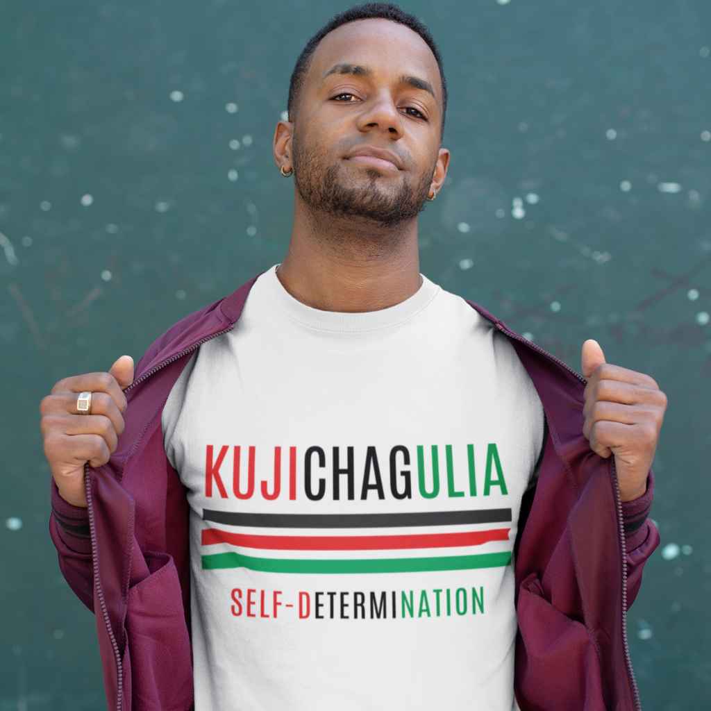 Young Black man wearing a white Kwanzaa T shirt that says Kujichagulia on it in Red, Black & Green.