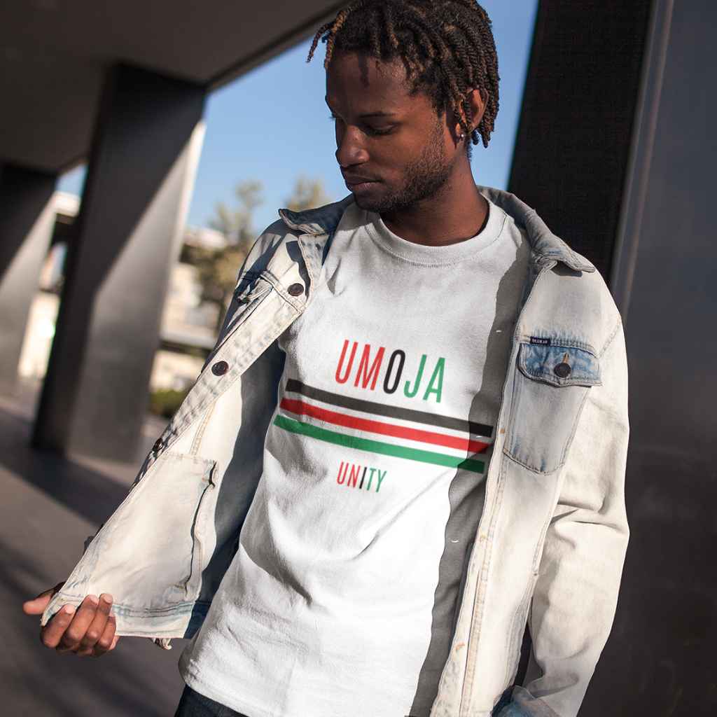 Young black man with locs, wearing a white Kwanzaa T Shirt that says Umoja in red, black and green, then there are Red Black & Green stripes and then Unity.
