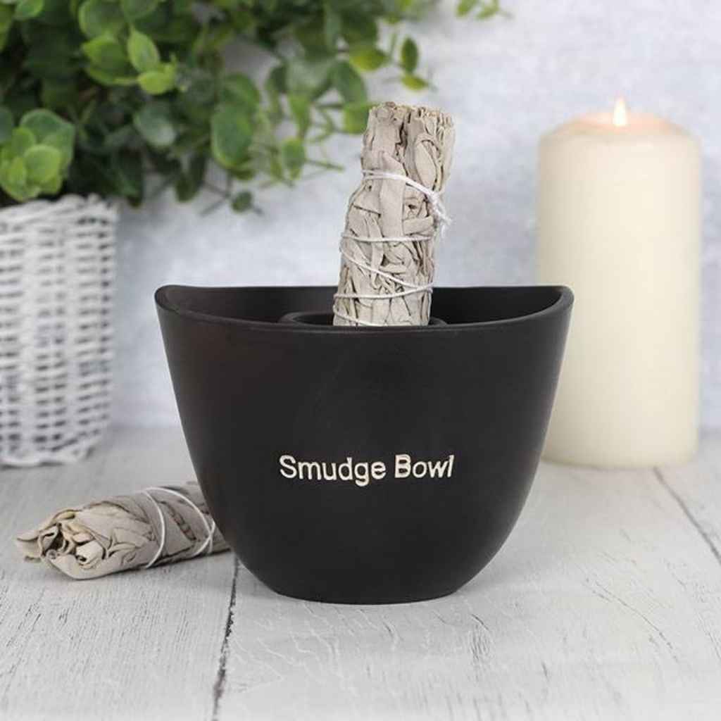 Smudge stick in a large black smudge bowl. The smudge stick is unlit and next to the bowl is another unlit smudge stick. The smudge bowl has the words Smudge Bowl etched into the front of the bowl. In the background is a lit candle to the right and a plant in a white wicker pot to the left.