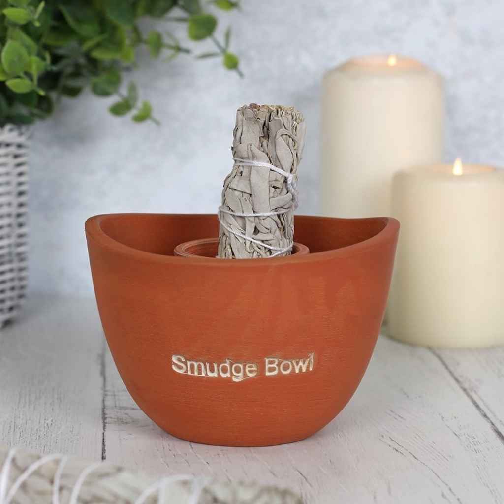 Large terracotta smudge bowl with a smudge stick showing at the top of the bowl. There are two lit candles behind the smudge bowl. The words smudge bowl are etched in white on the front of the smudge bowl.