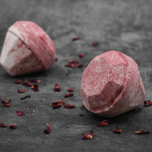 A picture of two luxurious looking pink diamond shaped bath bombs. The bath bombs are laying on their sides on a dark grey background. Sprinkles of dried red rose petals in the foreground and between the two bath bombs add to the sense of luxury.