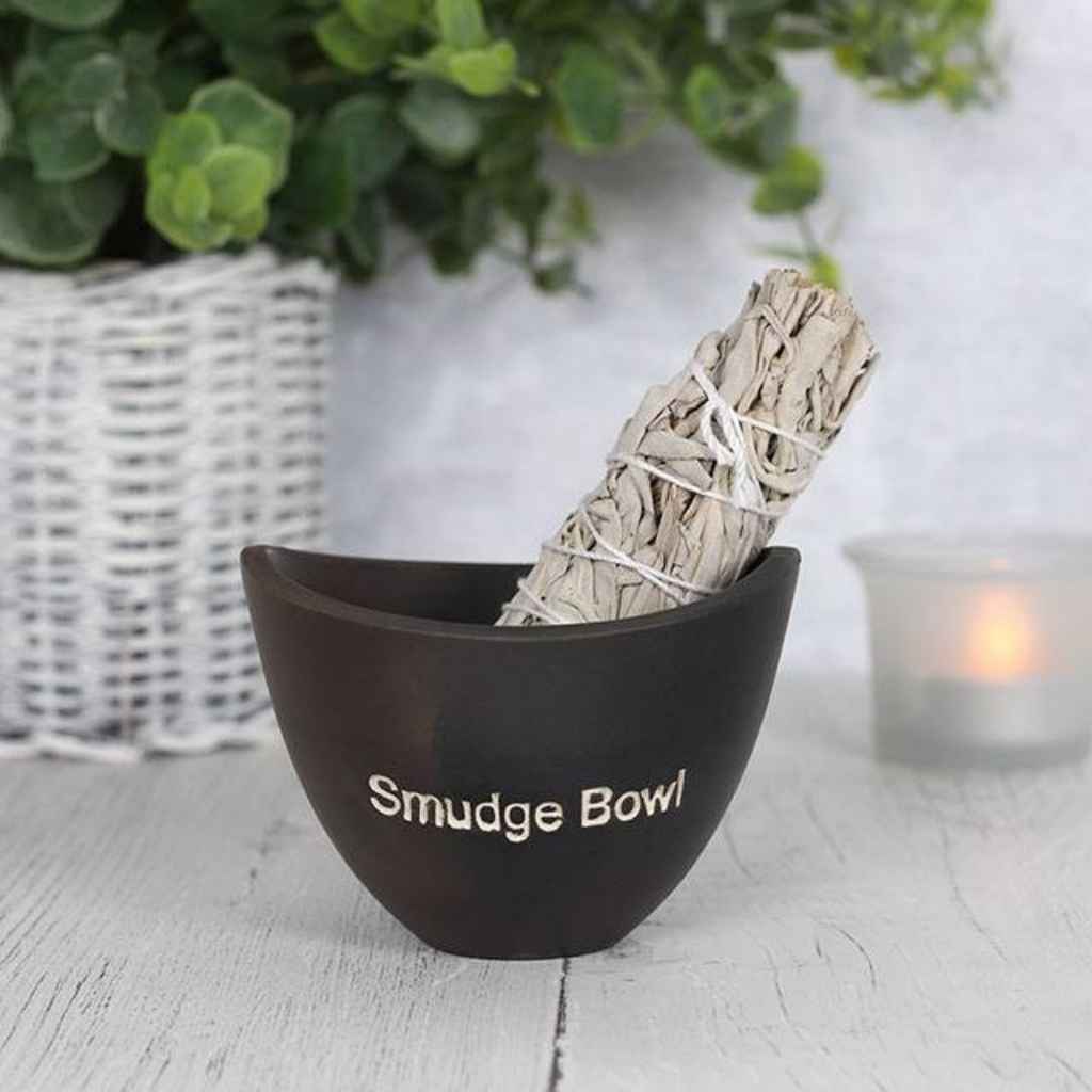 Small Black smudge bowl with a smudge stick showing at the top of the bowl. There is a lit candle behind the smudge bowl. The words smudge bowl are etched in white on the front of the smudge bowl.