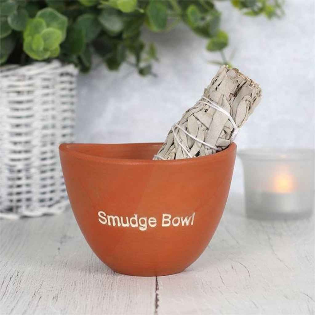 Small terracotta smudge bowl with a smudge stick showing at the top of the bowl. There is a lit candle behind the smudge bowl. The words smudge bowl are etched in white on the front of the smudge bowl.