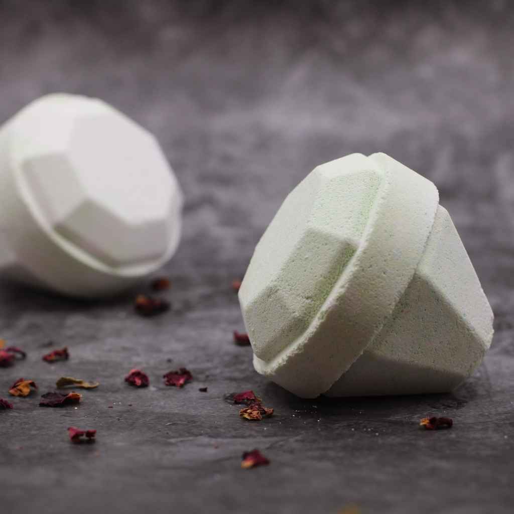 A picture of two luxurious looking light or pastel green diamond shaped bath bombs. The bath bombs are laying on their sides on a dark grey background. Sprinkles of dried red rose petals in the foreground and between the two bath bombs add to the sense of luxury.