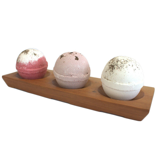 Wooden bath bomb holder on a white background. The bath bomb holder has three large bath bombs on it, one bath bomb in slightly dipped bay which is there to hold the bath bombs. Bath Bombs are pink with white, light pink and white. Each of the bath bombs in the bath bomb holder has some sort floral decoration on the top.