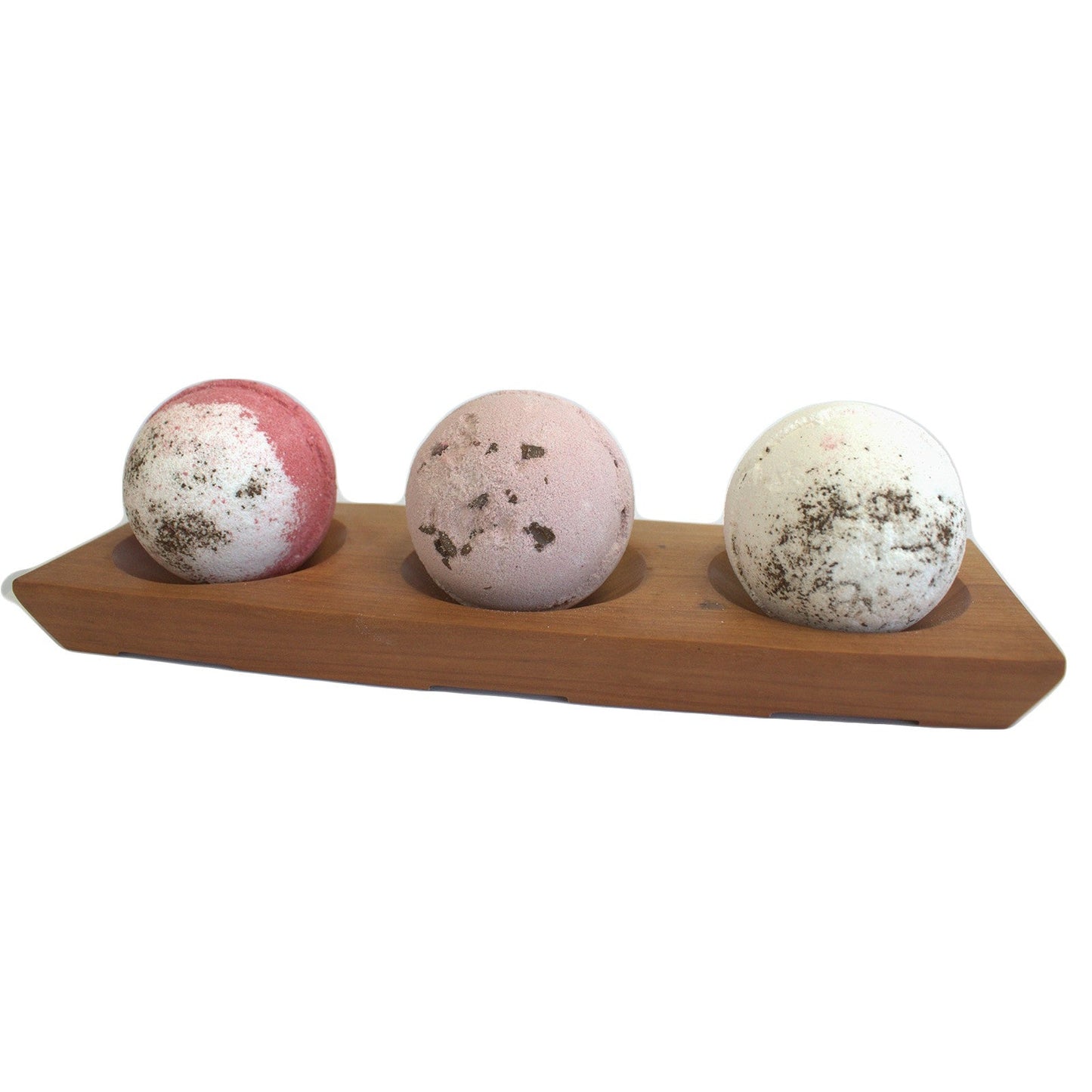 Wooden bath bomb holder on a white background. The bath bomb holder has three large bath bombs on it, one bath bomb in slightly dipped bay which is there to hold the bath bombs. Bath Bombs are pink with white, light pink and white. Each of the bath bombs in the bath bomb holder has some sort floral decoration on the top.